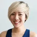 Smiling woman with short blonde hair wearing a blue sleeveless top, posed against a light background.