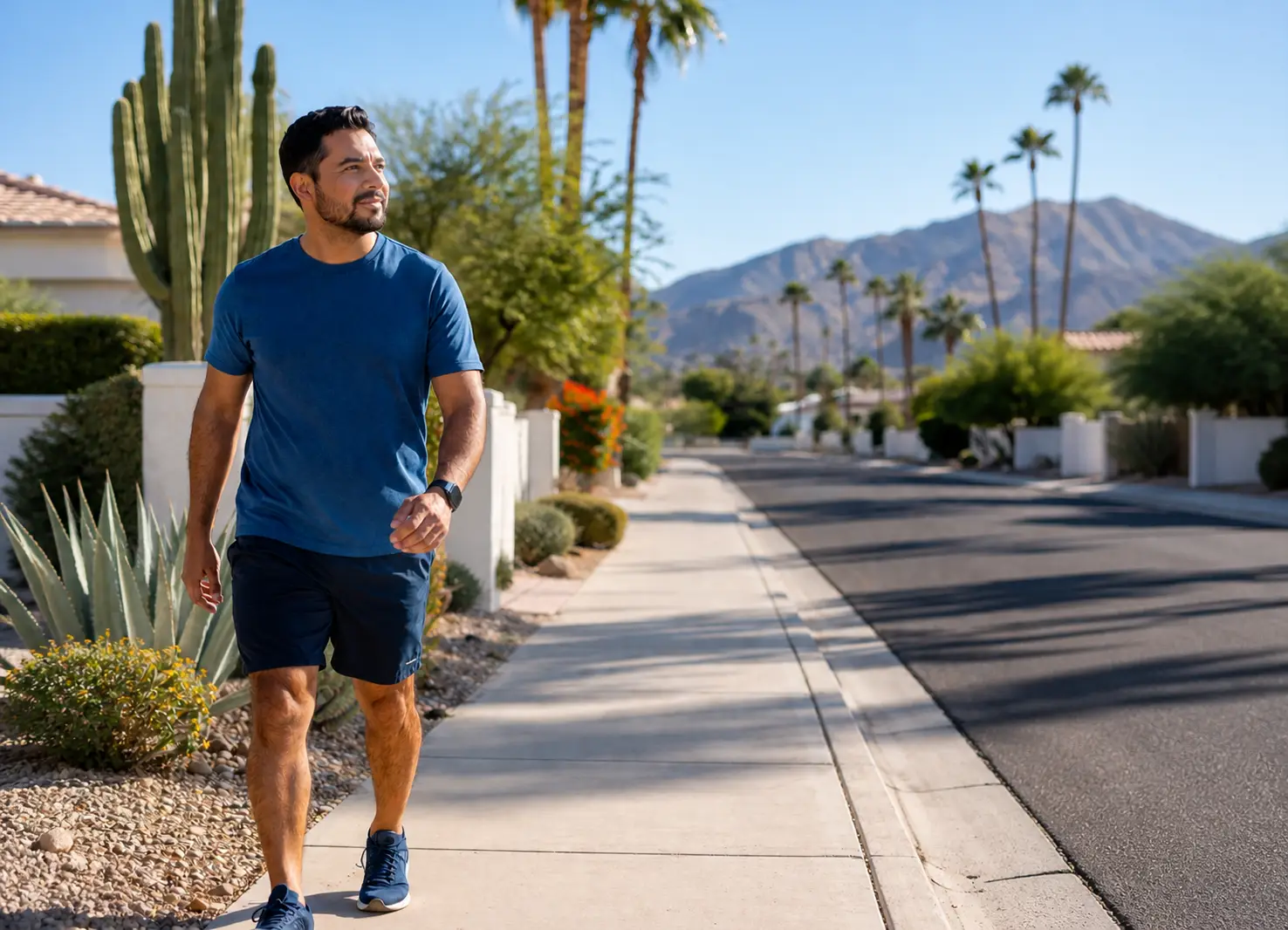 Man walking on a sunny desert suburb sidewalk in a blue T-shirt and dark shorts, with cacti, palm trees and mountains in the distance.