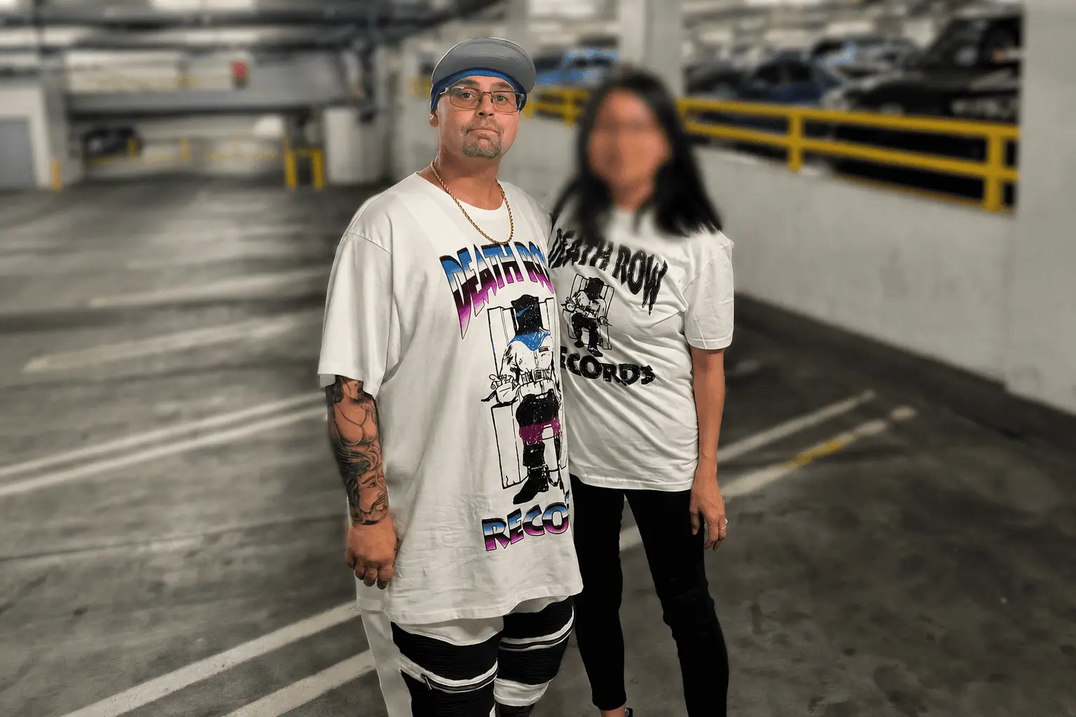 Two people pose in a parking garage, both wearing oversized white Death Row Records graphic tees; man in front with cap and tattoos.