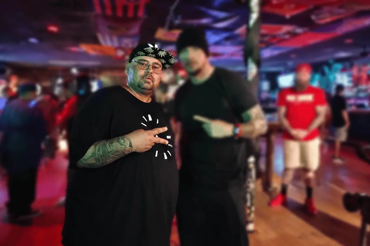Man with a tattooed arm in a black shirt and a patterned cap making a peace sign in a colorful, dimly lit nightclub. (Informative)