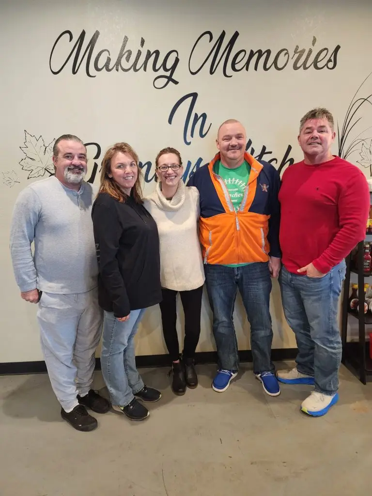 Five adults stand side by side indoors, smiling, in front of a wall with cursive text reading 'Making Memories'.