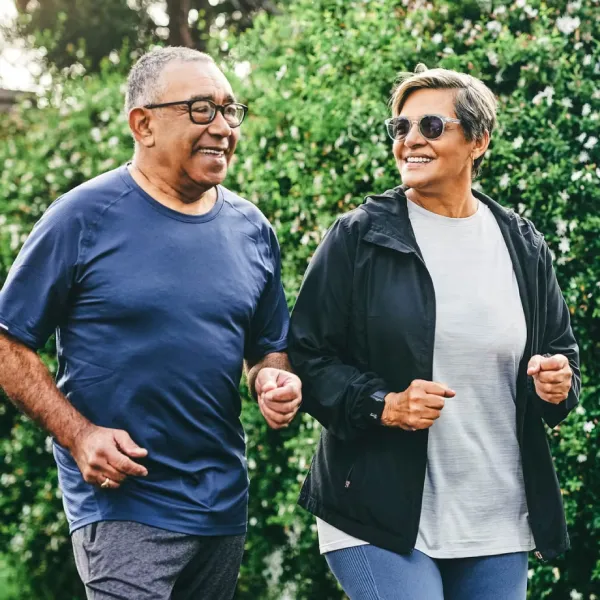 Two seniors jogging together outdoors, smiling and wearing athletic gear with a green leafy background.