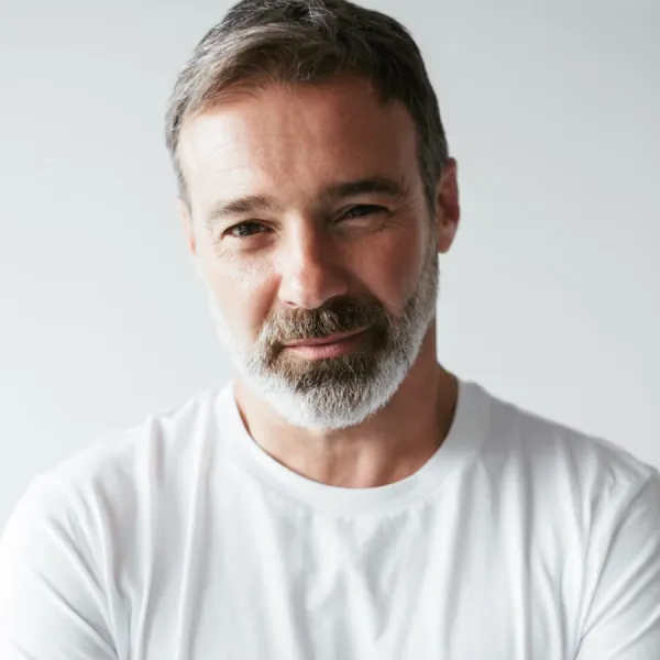 Portrait of a middle-aged man with salt-and-pepper hair and beard wearing a white T-shirt, looking at the camera with a gentle smile.
