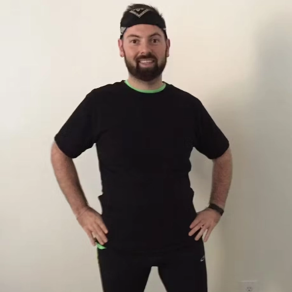 Bearded man in a black athletic shirt and headband stands with hands on hips against a light wall, looking at the camera.