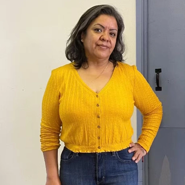 Woman standing indoors wearing a bright yellow cardigan and dark jeans, posing with her hand on her hip.