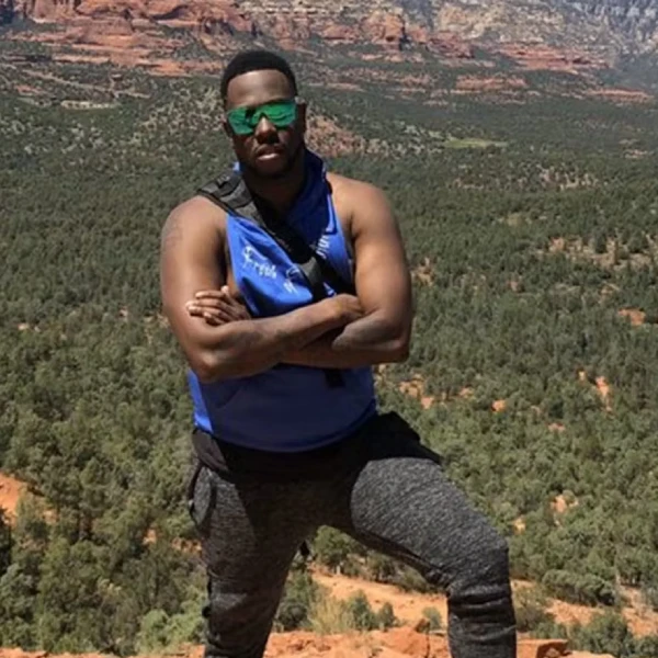 Man with crossed arms wearing a blue sleeveless shirt and green sunglasses on a canyon overlook.